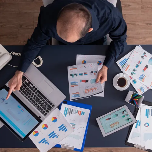 Top-down view of a professional analyzing charts and reports on a laptop, tablet, and printed documents spread across a workspace.