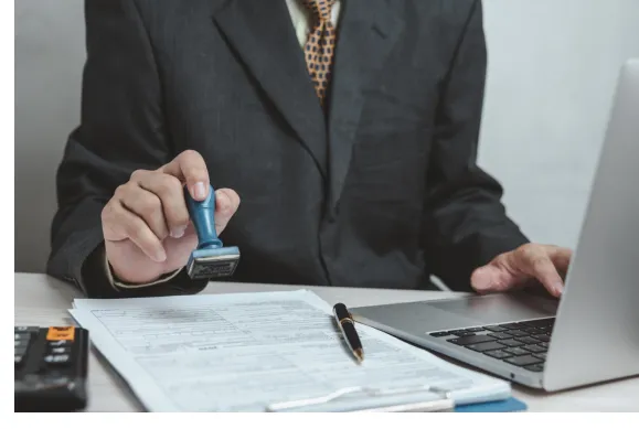 Business professional stamping official documents beside a laptop during an administrative review process.