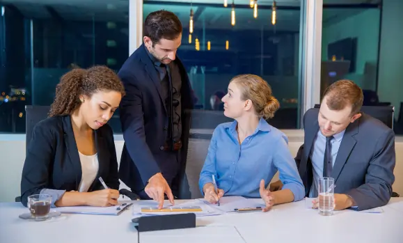 Four business professionals in formal attire reviewing charts and documents in a modern office during an evening Mainland Company Formation strategy session.