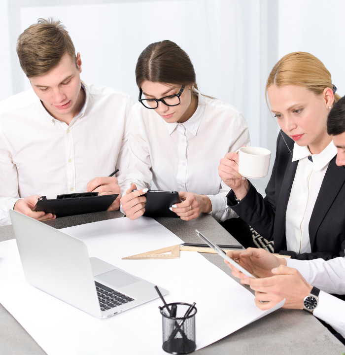 Office team focused on digital tasks, using tablets and a laptop while reviewing paperwork around a shared workspace.