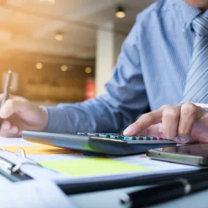 Individual recording calculations with a calculator and notepad next to an open laptop on an office table.