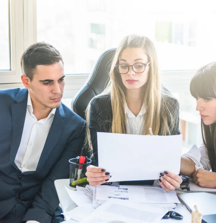Three office colleagues reviewing printed documents together at a desk during a workplace discussion.