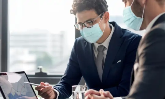 Two business professionals wearing face masks sitting at a desk, reviewing charts on a laptop and discussing financial or strategic data during a meeting.
